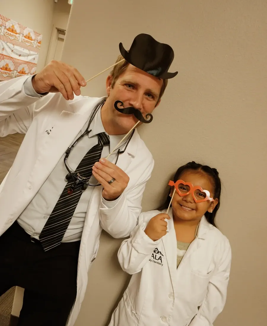 A dentist in a paper hat and moustache posing with a young patient in a lab coat.